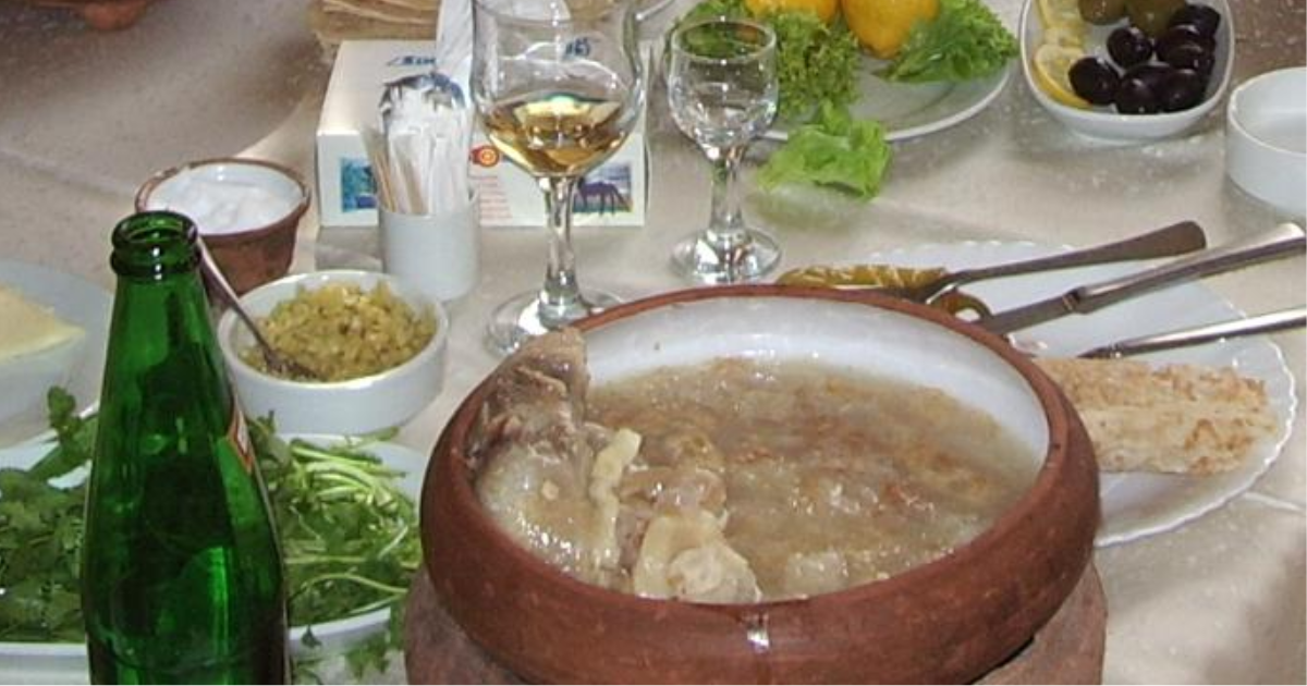 A bowl of Armenian khash — white, rich beef trotter broth garnished with dried lavash crumbles, served with garlic and fresh herbs on a wooden table