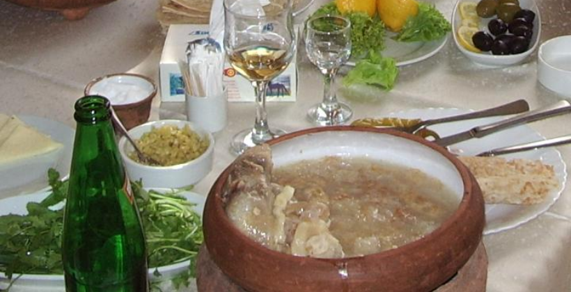 A bowl of Armenian khash — white, rich beef trotter broth garnished with dried lavash crumbles, served with garlic and fresh herbs on a wooden table